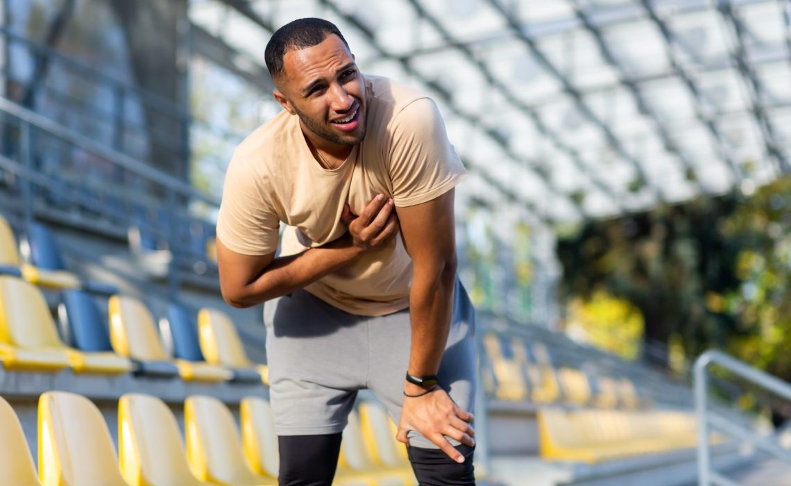Sick athlete after jogging and vigorous exercise has severe chest pain, heart attack in young afro american athlete, man holding hands on chest in stadium sunny afternoon.
